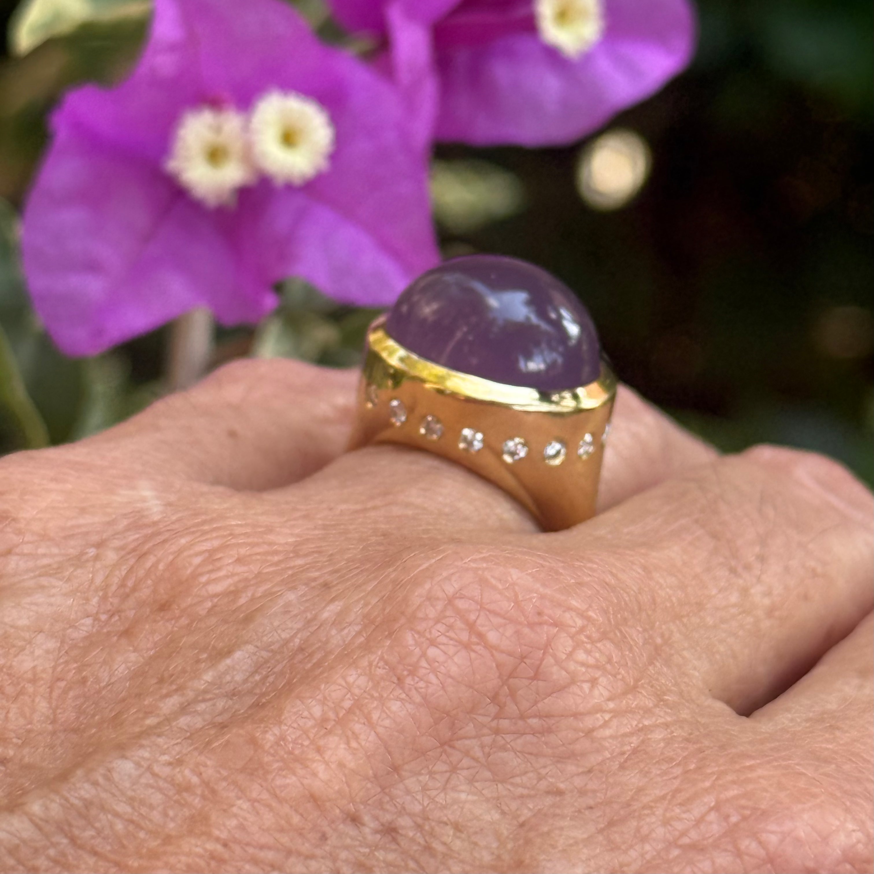 Gold ring with a purple gemstone on a hand, with purple flowers in the background- Ashleigh Branstetter